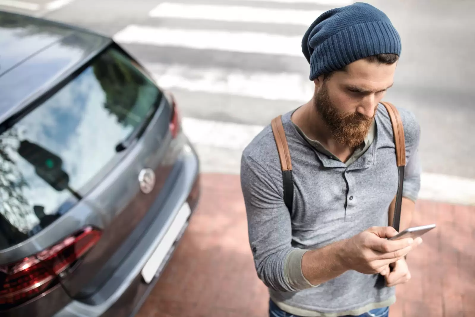 Ein Mann mit türkiser Mütze und Smartphone steht vor einem grauen Auto.