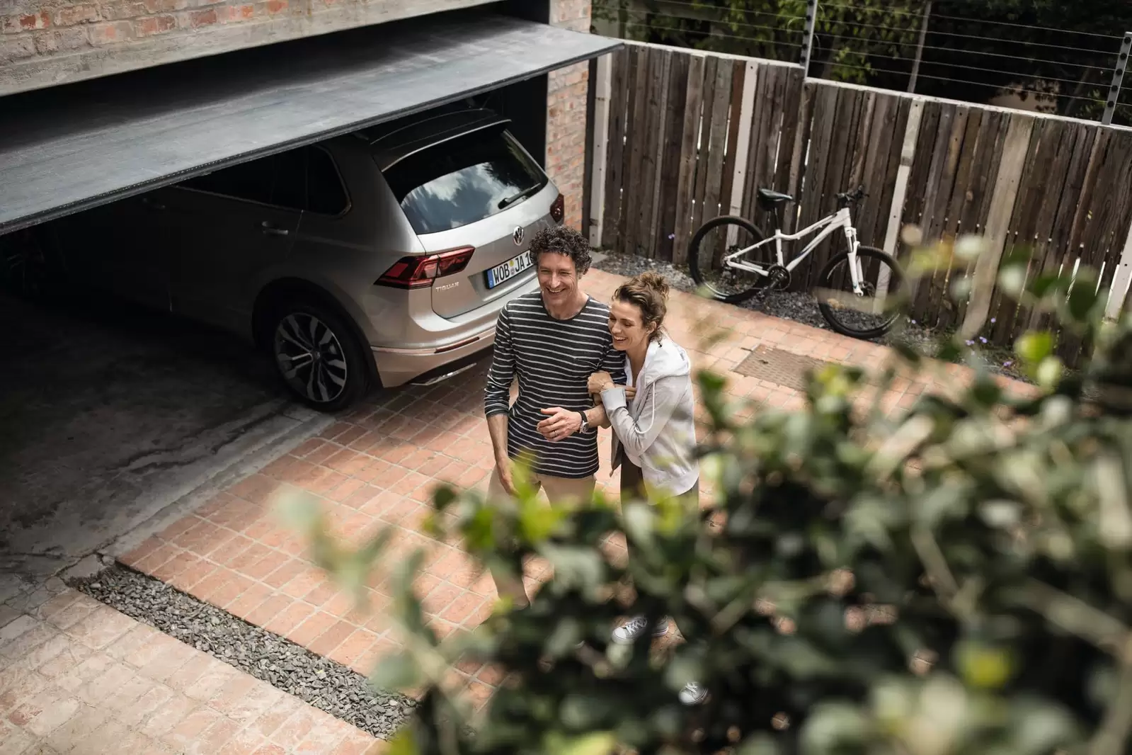 Ein Mann und eine Frau stehen lachend vor einem Auto auf der Einfahrt.